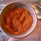 An earthenware bowl full of Rancho Gordo Sabor Vaquero chili poweder blend, sitting on a wooden table with a napkin, fork, and spoon to the right. 