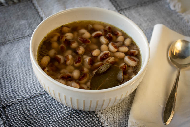 White bowl with cooked Rancho Gordo yelllow eye heirloom beans on a checkered cloth with a spoon resting on a napkin. 