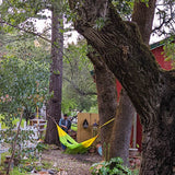 Handwoven Hammock with someone laying down and another person on the side with a red house in the back surronded with trees