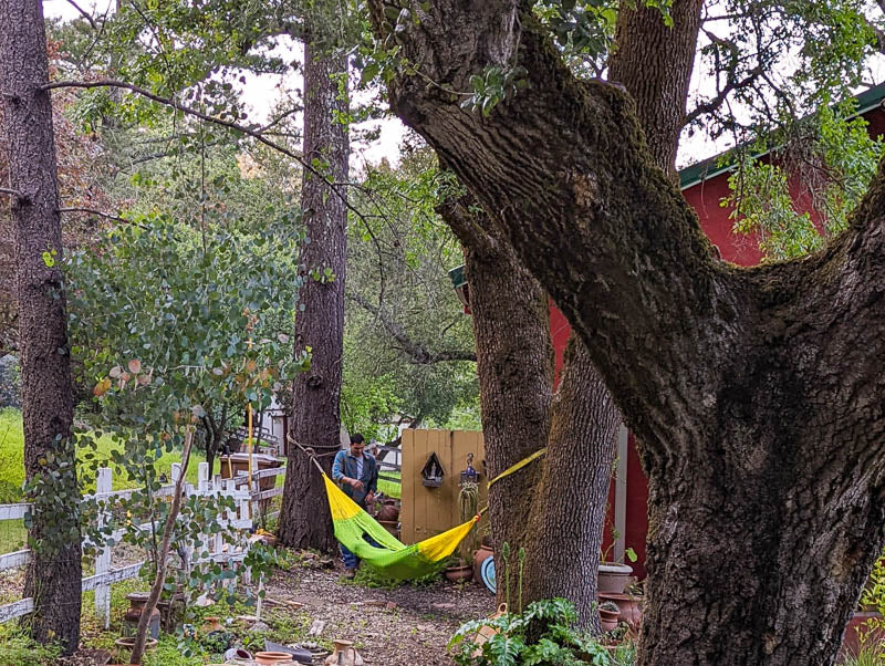 Handwoven Hammock with someone laying down and another person on the side with a red house in the back surronded with trees